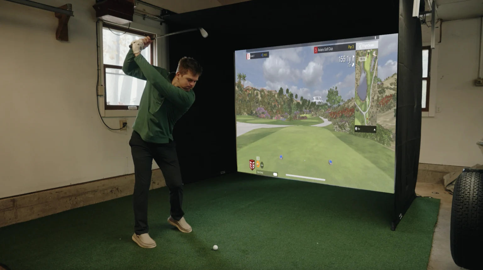 Person playing golf on a simulator in a room with a large screen displaying a virtual golf course.