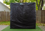 A black outdoor cover for a golf swing trainer, placed on a green mat with a wooden fence in the background.