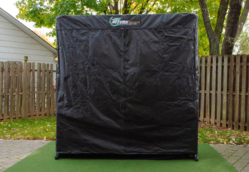 A black outdoor cover for a golf swing trainer, placed on a green mat with a wooden fence in the background.