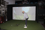 Person playing golf simulator indoors with a screen displaying a golf course.