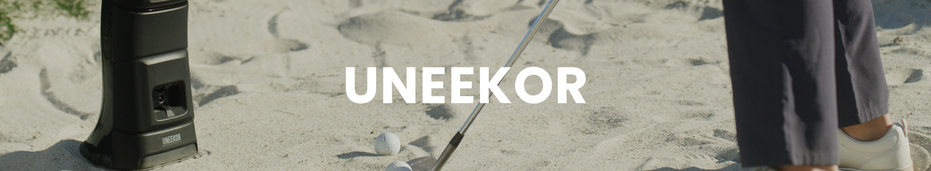 Person holding a walking stick on a sandy beach with 'UNEEEKOR' branding.
