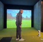 Man practicing golf swings in a simulator room with a screen displaying a golf course.