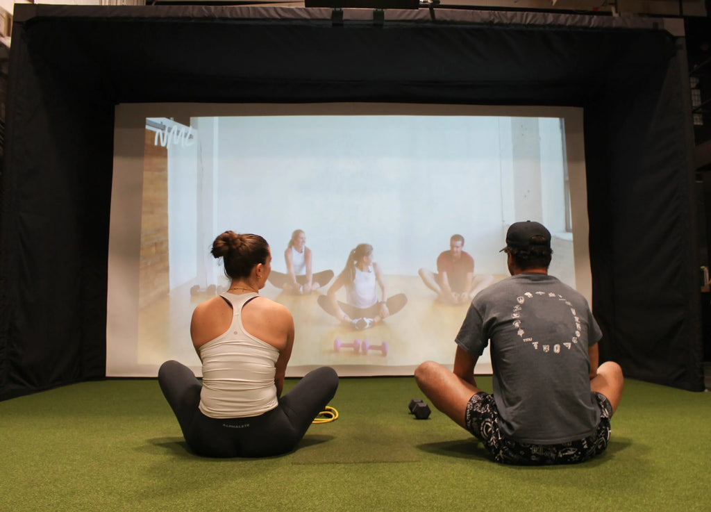 Two people sitting on the floor in front of a screen showing another person exercising.