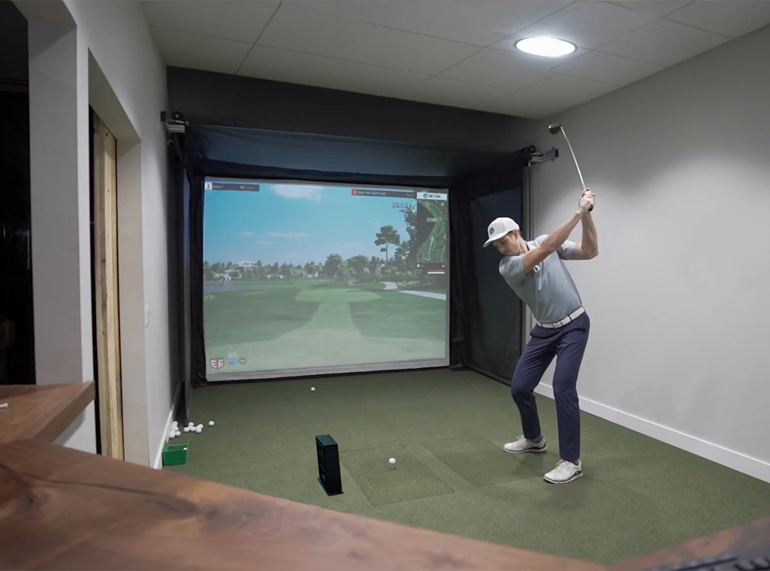 Person playing golf indoors using a simulator with a screen displaying a golf course.